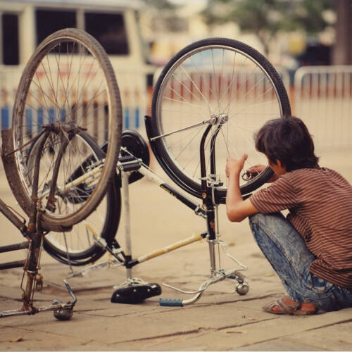 Photograph of a man fixing bikes. The bike nearest the man is dark blue/black while the bike to the left is deep red.